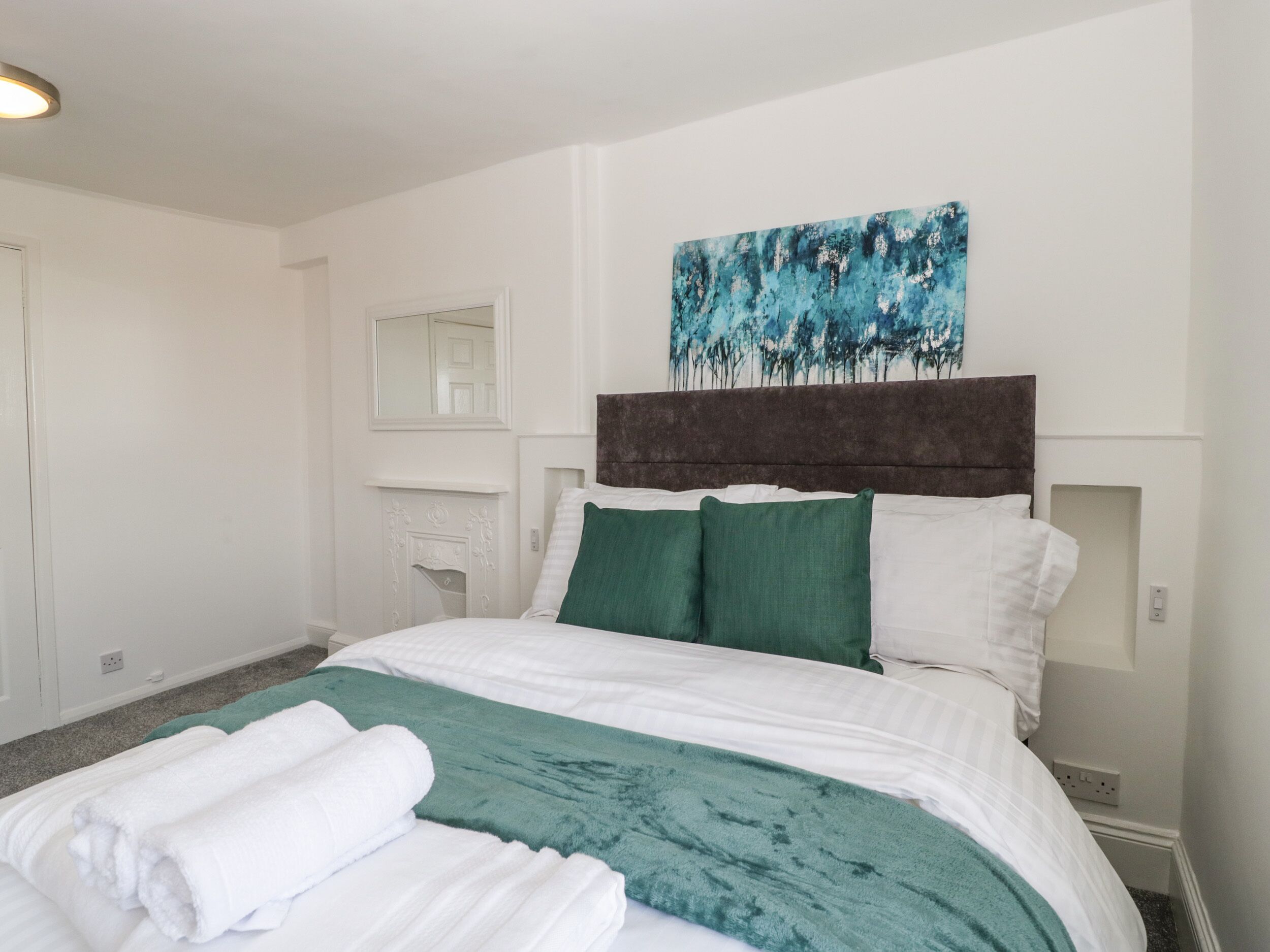 A hotel room with a large bed, white walls, a dark headboard, and a painting above the bed.