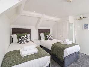 Une chambre d’hôtel moderne avec deux lits, chacun doté d’une tête de lit, de draps blancs et de coussins décoratifs verts. Une table de chevet blanche est placée entre les lits, et une porte avec un miroir et un tableau encadré se trouve sur le mur.