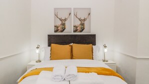 A neatly made bed with white linens and a yellow throw blanket, flanked by two white pillows and a dark headboard. Two wall-mounted lamps are on either side of the bed. - Alcombe House (Minehead)