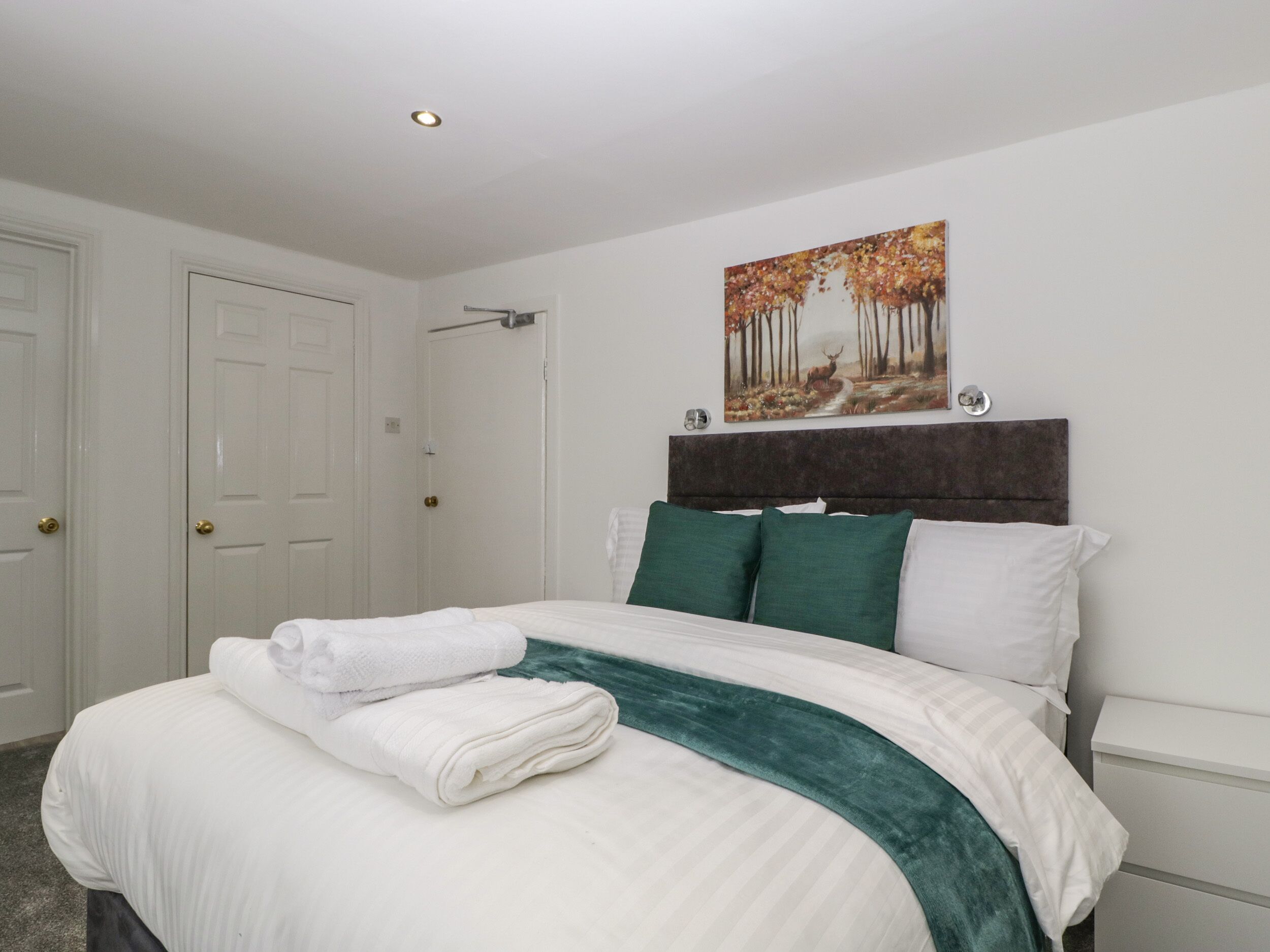 A neatly made bed with white linens and a green throw blanket, a painting of a forest scene on the wall, and a white nightstand with a drawer.