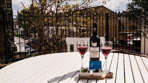 Uma mesa com uma garrafa de vinho Chianti Classico e dois copos sobre uma toalha de mesa branca, com um portão de metal e árvores ao fundo.