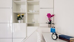 Um quarto branco com uma mesa, uma estante de livros, um relógio e duas plantas em vasos.