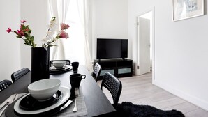 Uma sala de estar moderna com uma mesa de jantar posta para dois, uma televisão de tela plana e uma janela grande com cortinas finas.