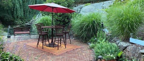 Un patio con una mesa y sillas de ladrillo, rodeado de exuberante follaje y un paraguas rojo.