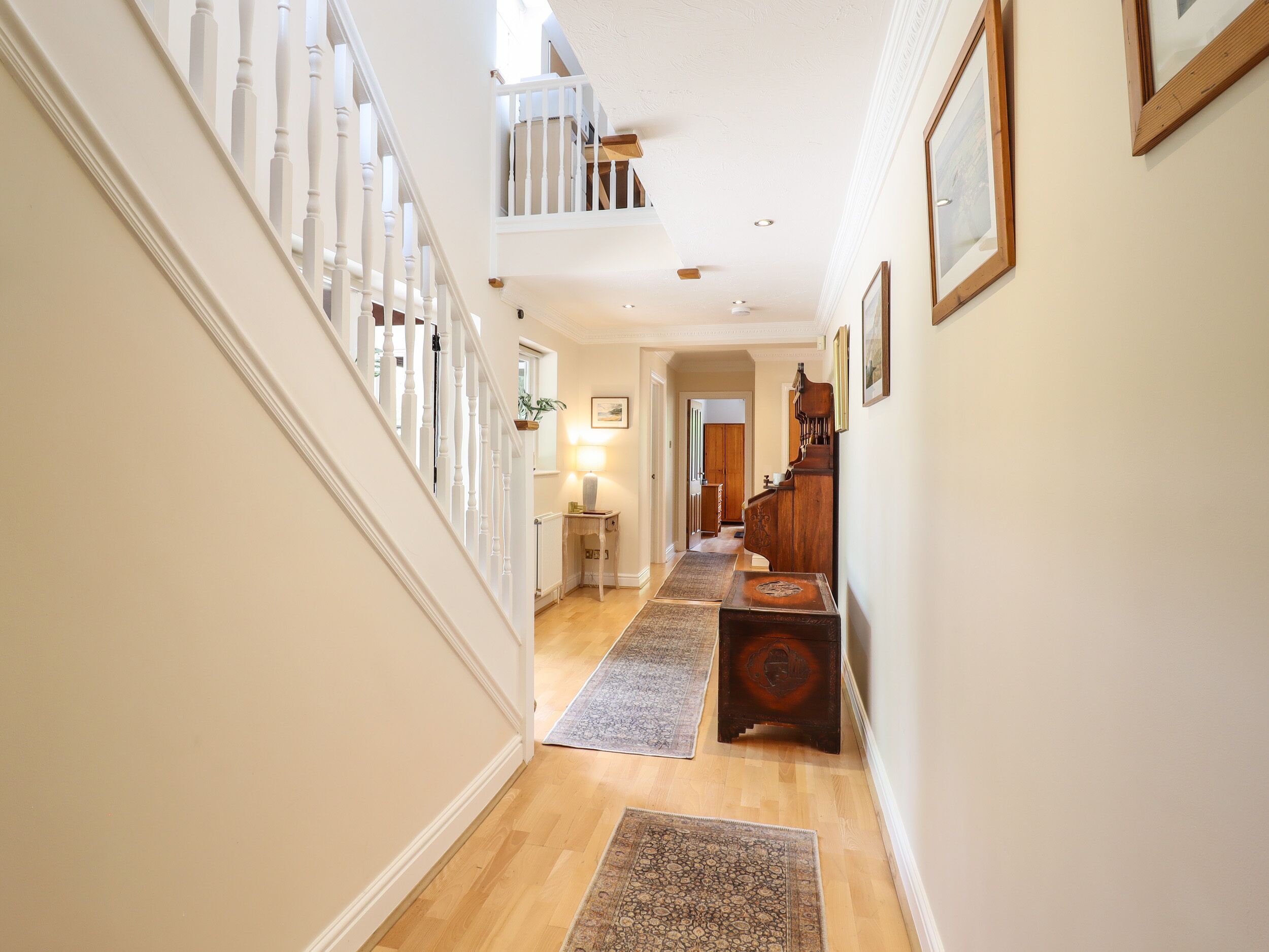 A hallway with a wooden floor, white walls, and a staircase with white balusters. There are two rugs on the floor and a wooden bench with a decorative backrest.