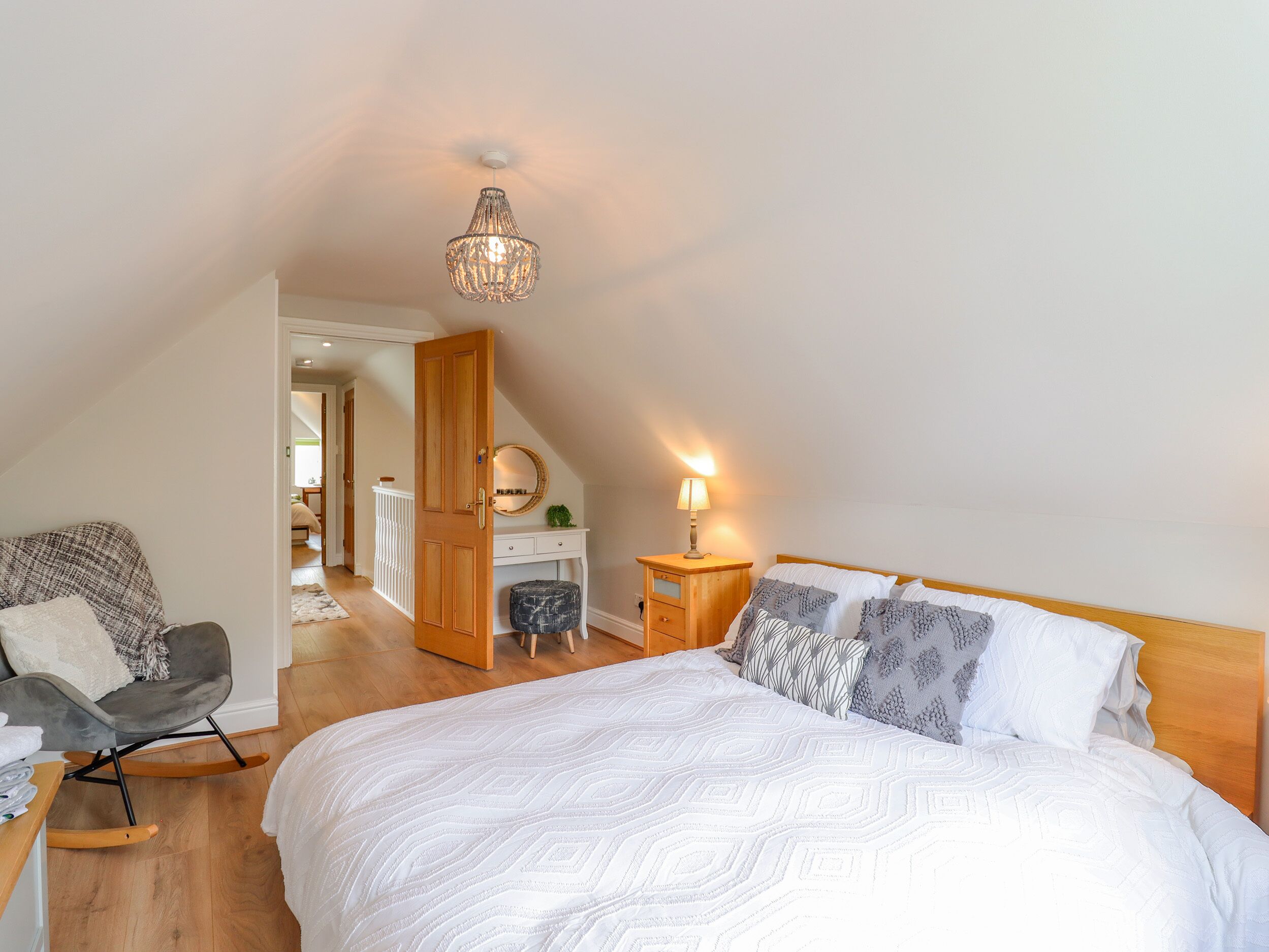 A bedroom with a large bed, a chair, a desk, a mirror, and a hanging light fixture.