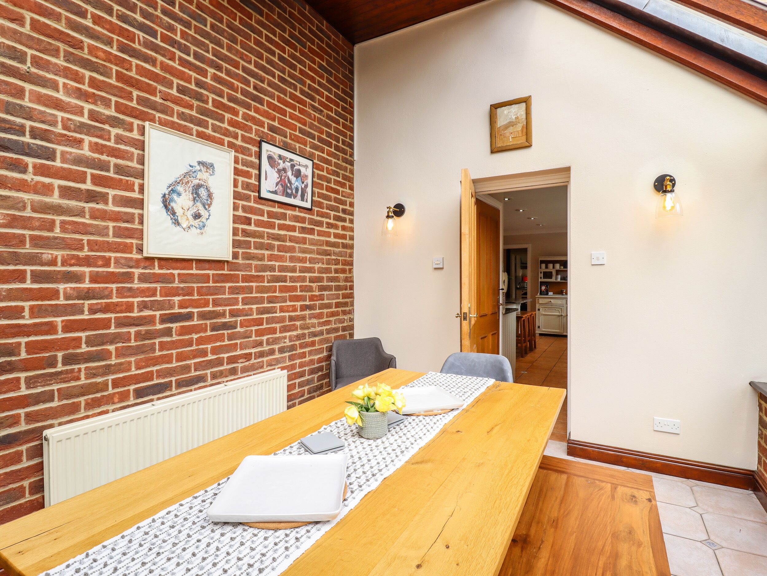 A dining area with a wooden table, chairs, and a radiator. There are framed pictures on the brick wall and a door leading to another room.