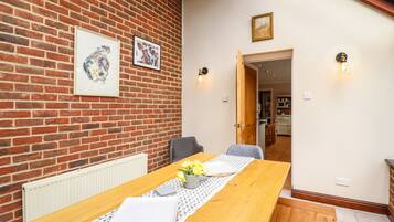 A dining area with a wooden table, chairs, and a radiator. There are framed pictures on the brick wall and a door leading to another room.