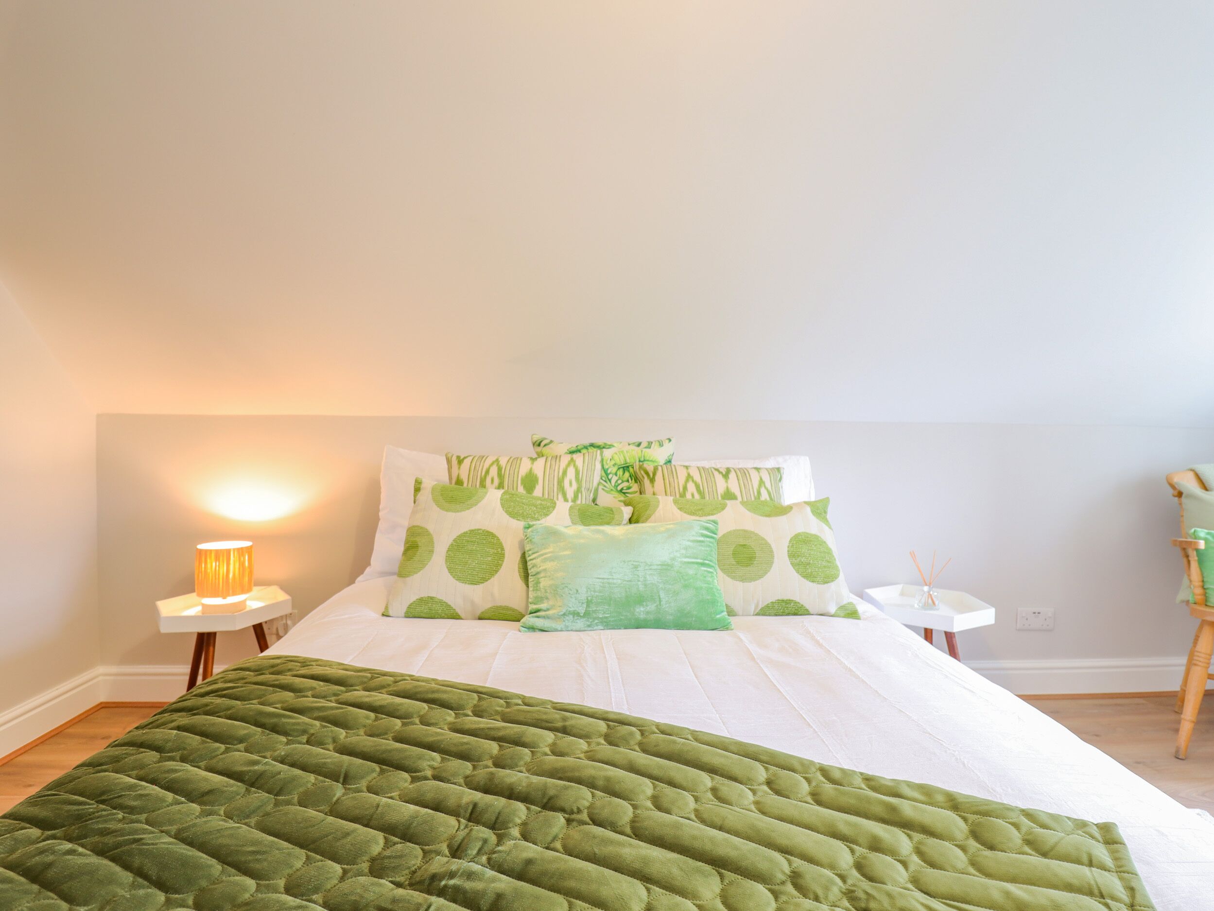 A neatly made bed with green bedding and a quilted green duvet, a bedside lamp, and a small white table with a green plant.