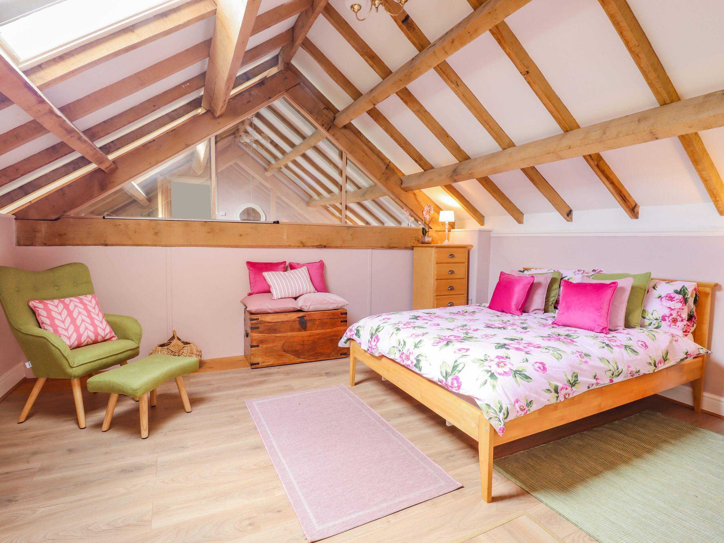 Un dormitorio con techo inclinado, vigas de madera, una cama con ropa de cama floral, un sillón verde, un cofre de madera y un pequeño banco.