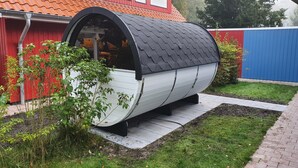 Una estructura petita i tancada, similar a un vehicle, amb un sostre corbat, situada en una zona de jardí amb un edifici vermell al fons.