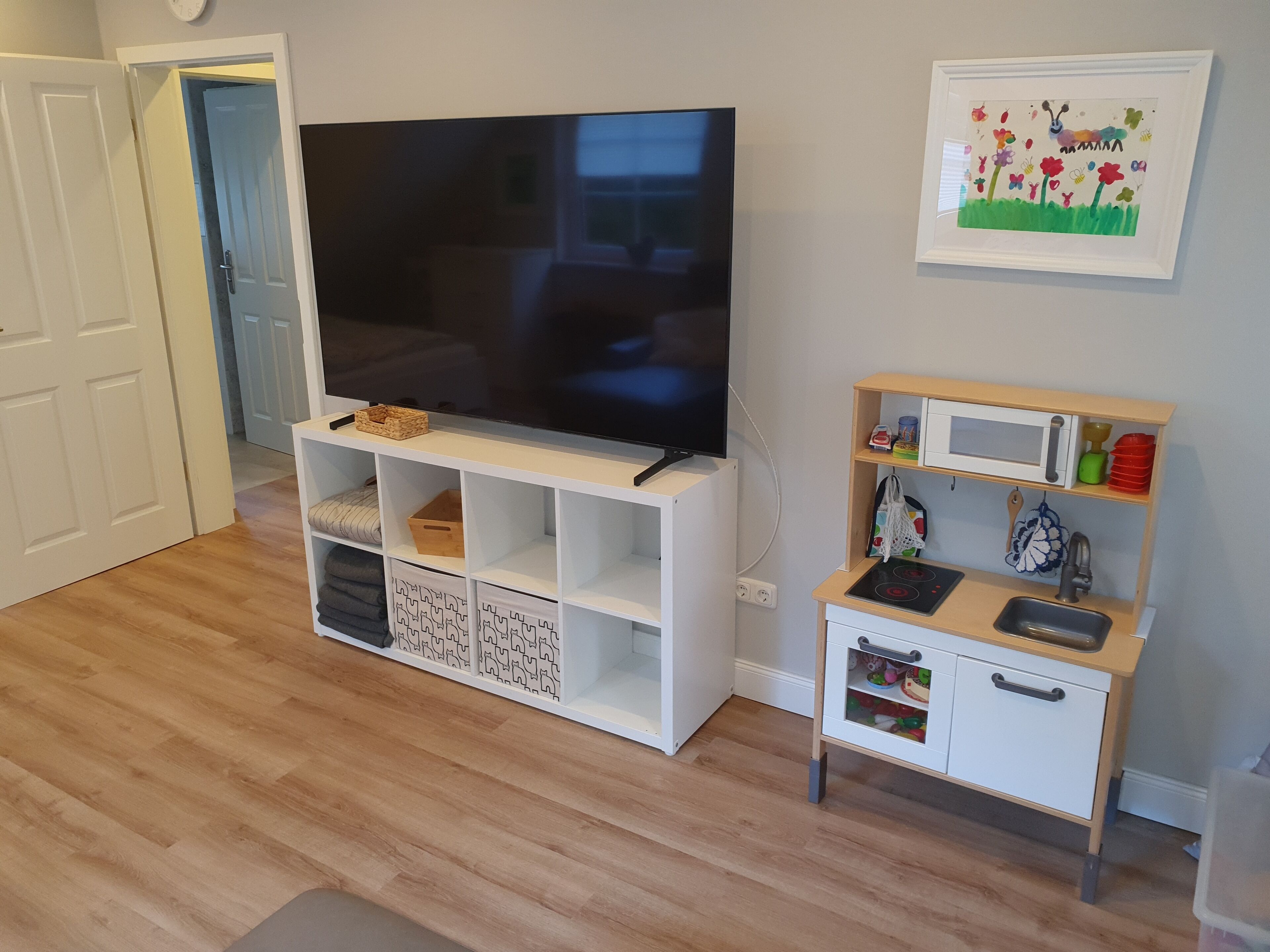 Ein Wohnzimmer mit Fernseher auf einer weißen Unterhaltungsanlage, eine kleine Küchenzeile mit Mikrowelle und Spüle, und eine Tür zu einem weiteren Raum.
