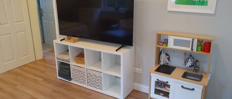 Ein Wohnzimmer mit Fernseher auf einer weißen Unterhaltungsanlage, eine kleine Küchenzeile mit Mikrowelle und Spüle, und eine Tür zu einem weiteren Raum.