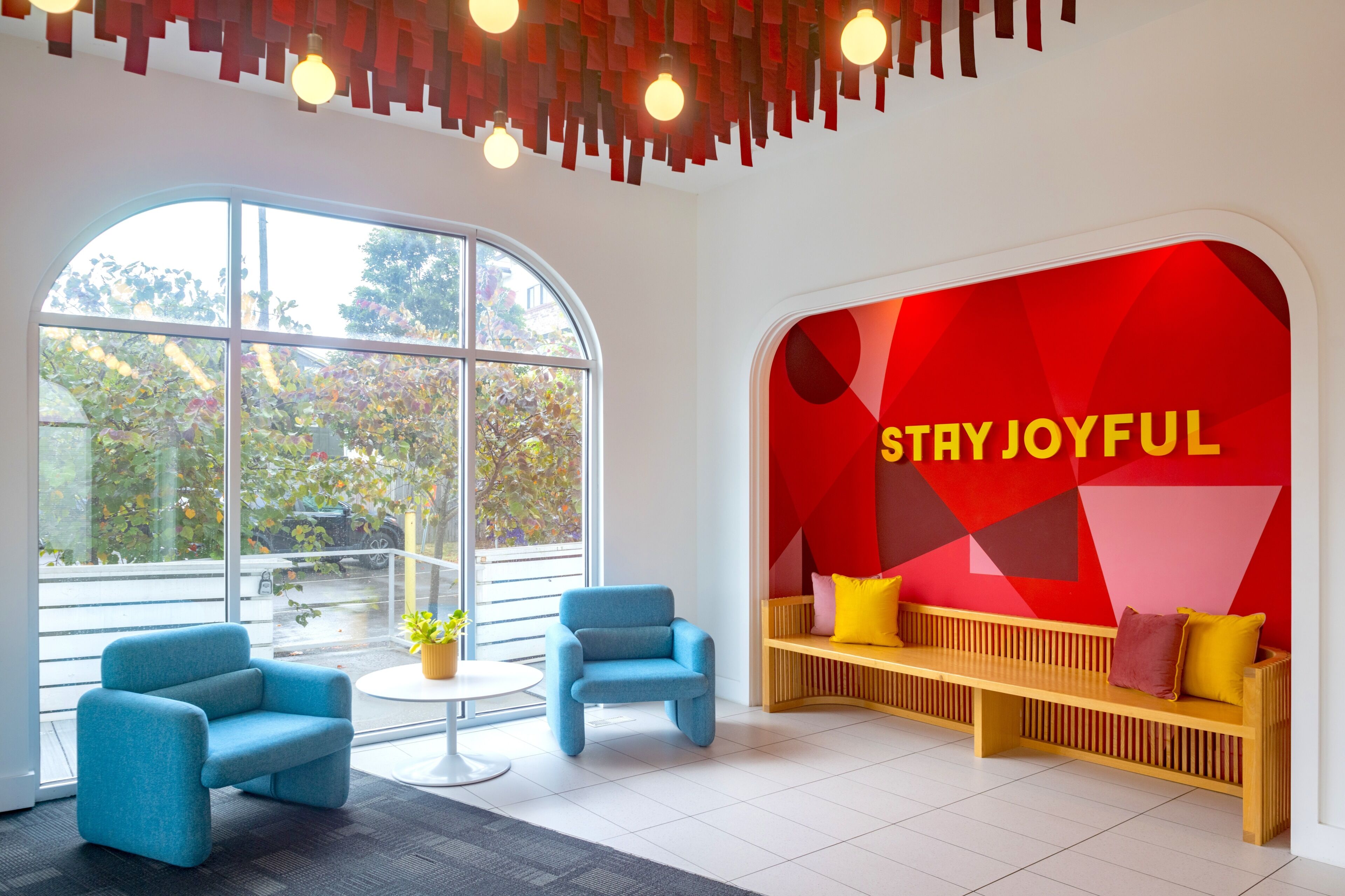 Un interior moderno con un gran ventanal, dos sillones azules, una mesa redonda, un banco de madera con cojines y una pared roja con el texto 'STAY JOYFUL'.