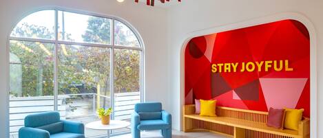 Un interior moderno con un gran ventanal, dos sillones azules, una mesa redonda, un banco de madera con cojines y una pared roja con el texto 'STAY JOYFUL'.