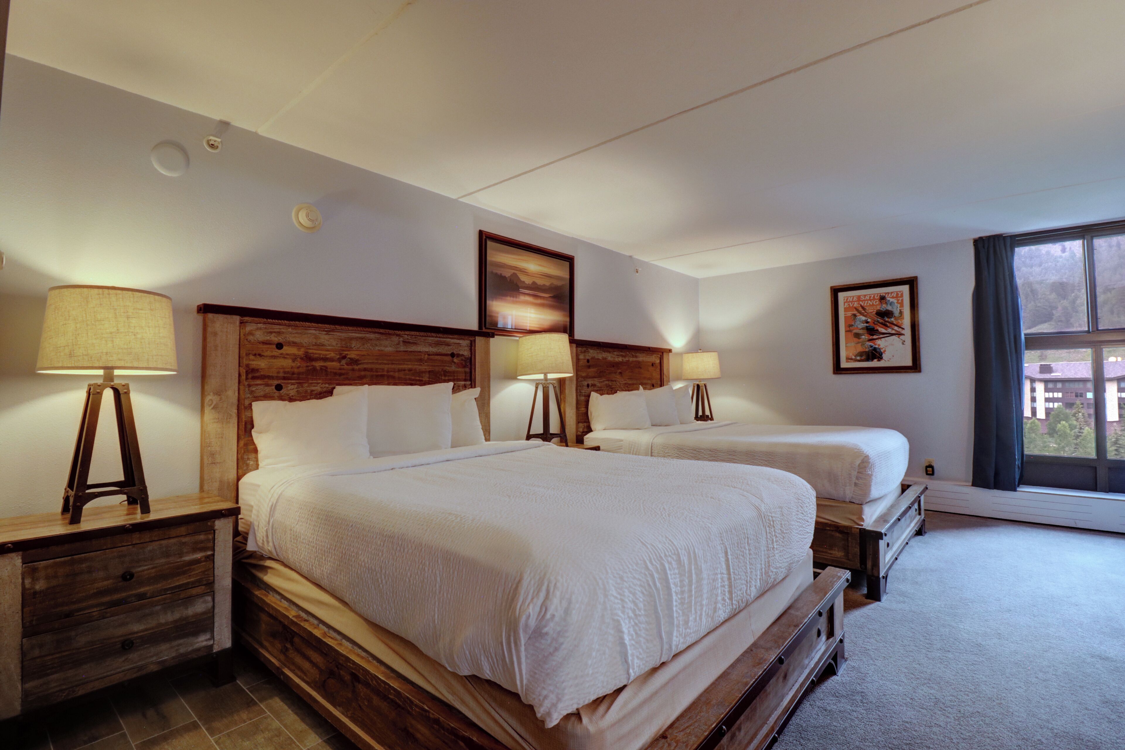 Habitación de hotel con dos camas, cabeceros de madera, lámparas de noche y un ventanal con vistas.