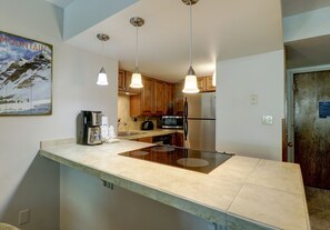 A modern kitchen with wooden cabinets, a stainless steel refrigerator, a built-in island with a glass top, pendant lighting, and a coffee maker.