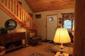 A cozy living room with a wooden staircase, a comfortable armchair, a lamp, and a door with a heart-shaped decoration. - Pond side cabin nestled in the woods (Lakeside)