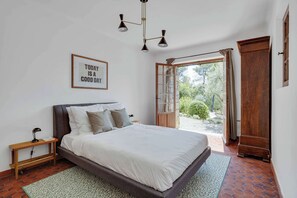A bedroom with a bed, a nightstand, a framed quote on the wall, a wooden wardrobe, and a view through an open door.