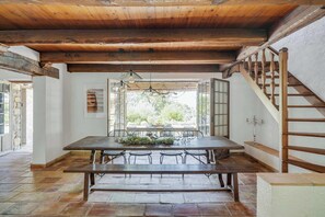 A rustic dining area with a wooden table and chairs, a staircase, and a view of greenery outside.