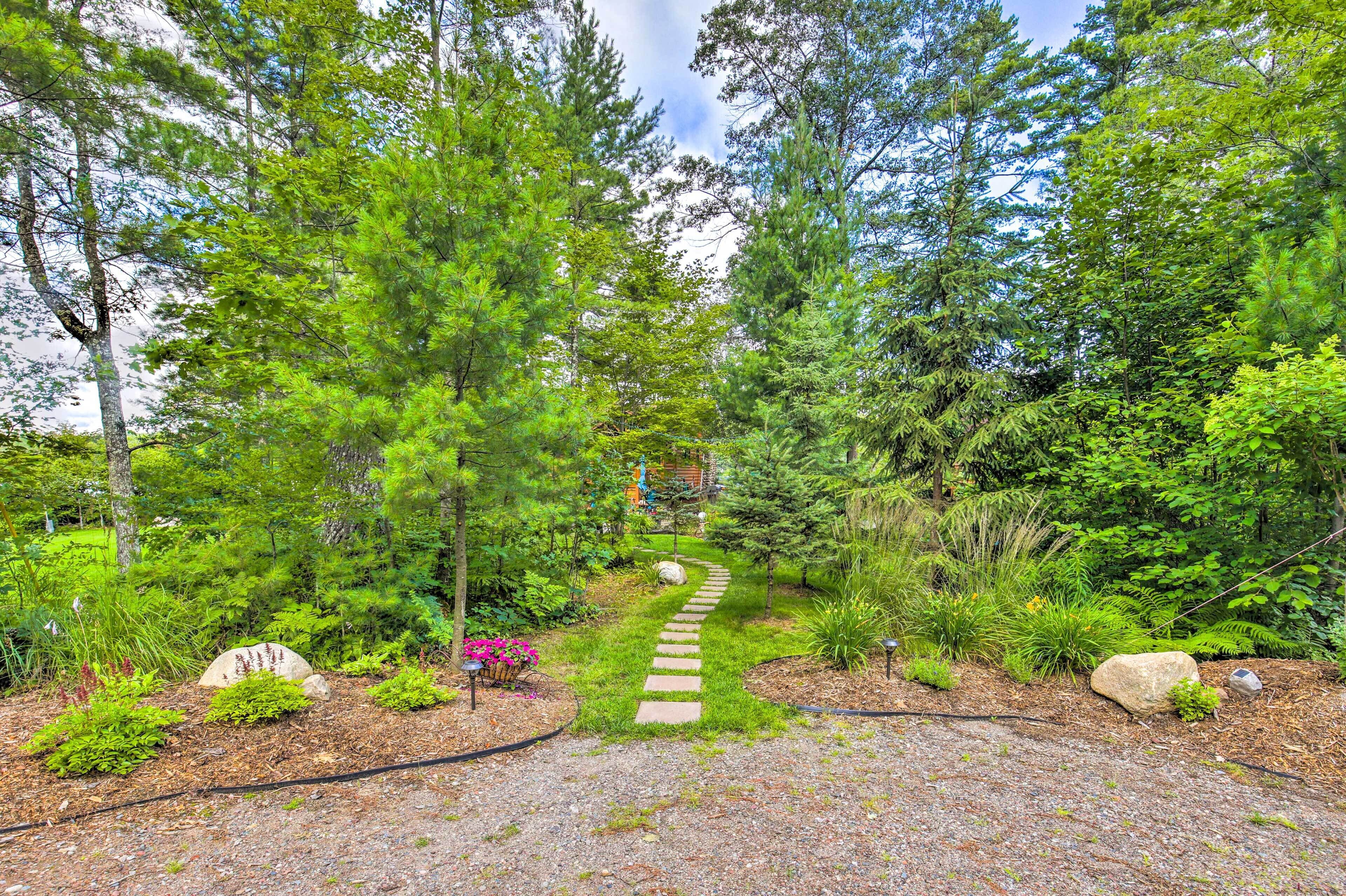 A garden pathway leading through a forested area with a stone path, surrounded by various plants and trees.