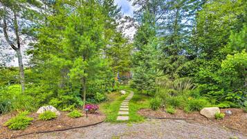 A garden pathway leading through a forested area with a stone path, surrounded by various plants and trees.