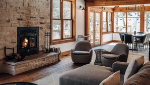 Una sala de estar acogedora con chimenea de piedra, techo de madera y ventanas amplias con vistas a un paisaje nevado.