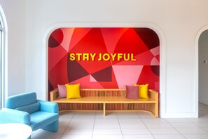 Un interior moderno con un banco y una silla azul, una pared con un diseño geométrico y el texto 'STAY JOYFUL' en la pared.