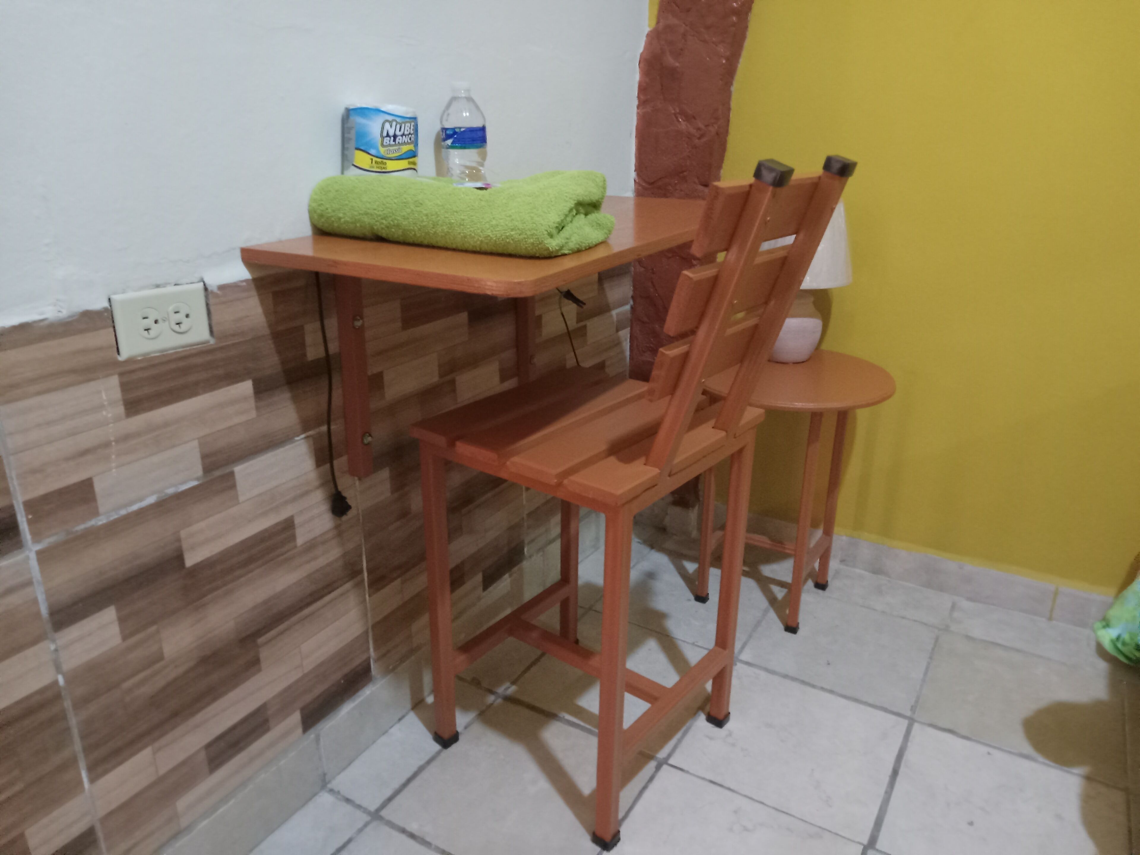 Una habitación pequeña con una mesa y silla de madera, una toalla verde, una botella de agua y una caja de pañuelos.
