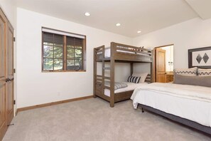 Quarto com uma cama de solteiro, uma cama beliche, uma janela com persianas, uma mesinha de cabeceira e um quadro na parede.