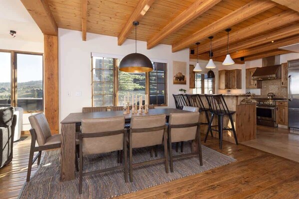 A spacious dining area with a wooden table and chairs, a large window with a view, and a modern kitchen with stainless steel appliances. - Ski-In/Ski-Out Big Sky Home with Incredible Views (Big Sky)
