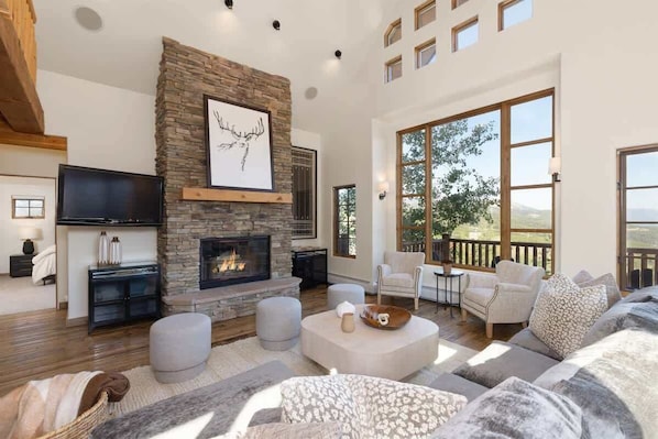 A spacious living room with a stone fireplace, comfortable seating, and large windows offering a view of the outdoors.