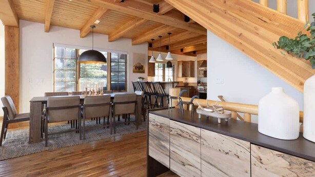 A spacious kitchen with wooden beams, a dining area with a large table and chairs, and a view of the outdoors through a window. - Ski-In/Ski-Out Big Sky Home with Incredible Views (Big Sky)