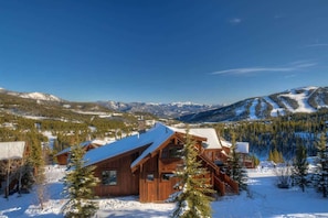 A wooden cabin with a sloped roof surrounded by snow-covered trees and mountains in the background.