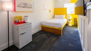 A hotel room with a bed, a desk with a lamp, a small cabinet, and a painting on the wall.