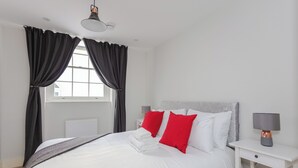 A neatly made bed with white bedding and red pillows, a bedside table with a lamp, and a window with black curtains.