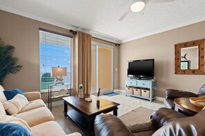 A living room with a sofa, a coffee table, a television, and a sliding glass door leading to a balcony. - Treasure Island 1806 (Panama City Beach)