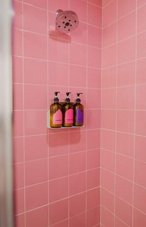 A bathroom with pink tiled walls and a showerhead. There are three bottles of shampoo and conditioner on a shelf. - The Gallatin (211) East Nashville Boutique Hotel (Nashville)