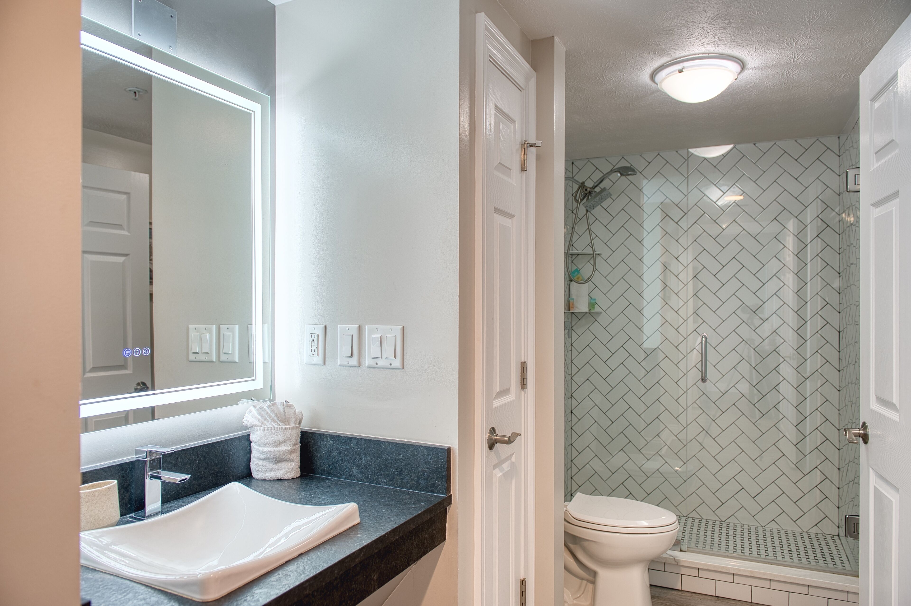A bathroom with a white sink, mirror, and toilet. There is a shower area with a glass door and a herringbone tile pattern on the walls.