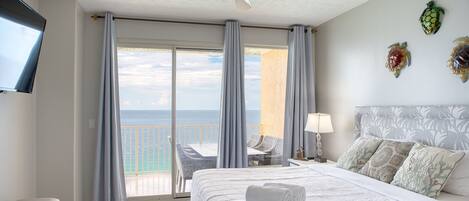 Habitación de hotel con cama, ventilador de techo, televisión y balcón con vista al océano, además de detalles decorativos en la pared.