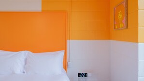 Una habitació d'hotel amb una combinació de colors taronja i blanca, un llit amb roba de llit blanca, un rellotge digital i una fotografia emmarcada a la paret.