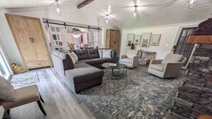 Un salon spacieux avec un grand canapé d’angle, deux fauteuils, une table basse et un tapis à motifs. On y trouve également une armoire en bois et un mur décoratif en pierre.