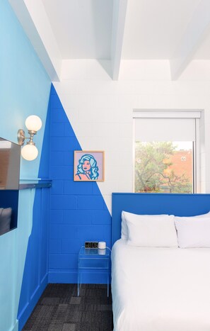 A room with a blue accent wall, a bed with white bedding, a small bedside table with a clock, a framed picture, and a window with a view of trees.