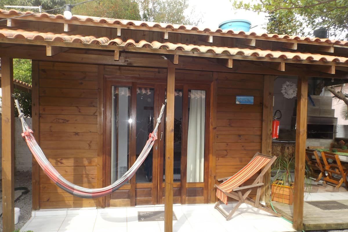 Uma cabana de madeira com telhado de telhas, uma rede, uma cadeira de praia listrada e uma pequena mesa com uma planta.