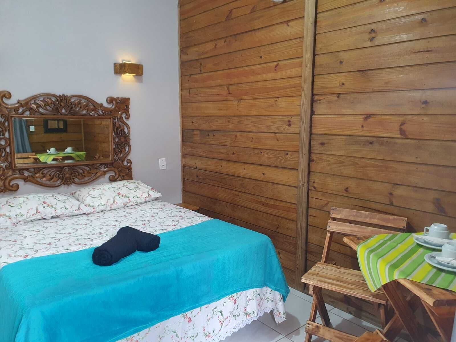 A bedroom with a wooden wall, a bed with floral bedding, a mirror, and a small table with a striped tablecloth.