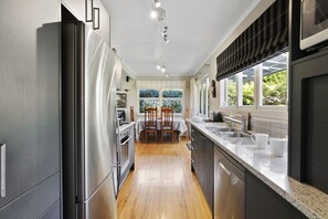 A modern kitchen with stainless steel appliances, a wooden dining table, and a large window providing a view of greenery. - Pukeko Lodge - Tūrangi Holiday Home (Turangi)