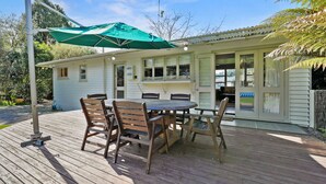 A wooden deck with a table and chairs, a green umbrella, and a building with large windows and a door. - Pukeko Lodge - Tūrangi Holiday Home (Turangi)