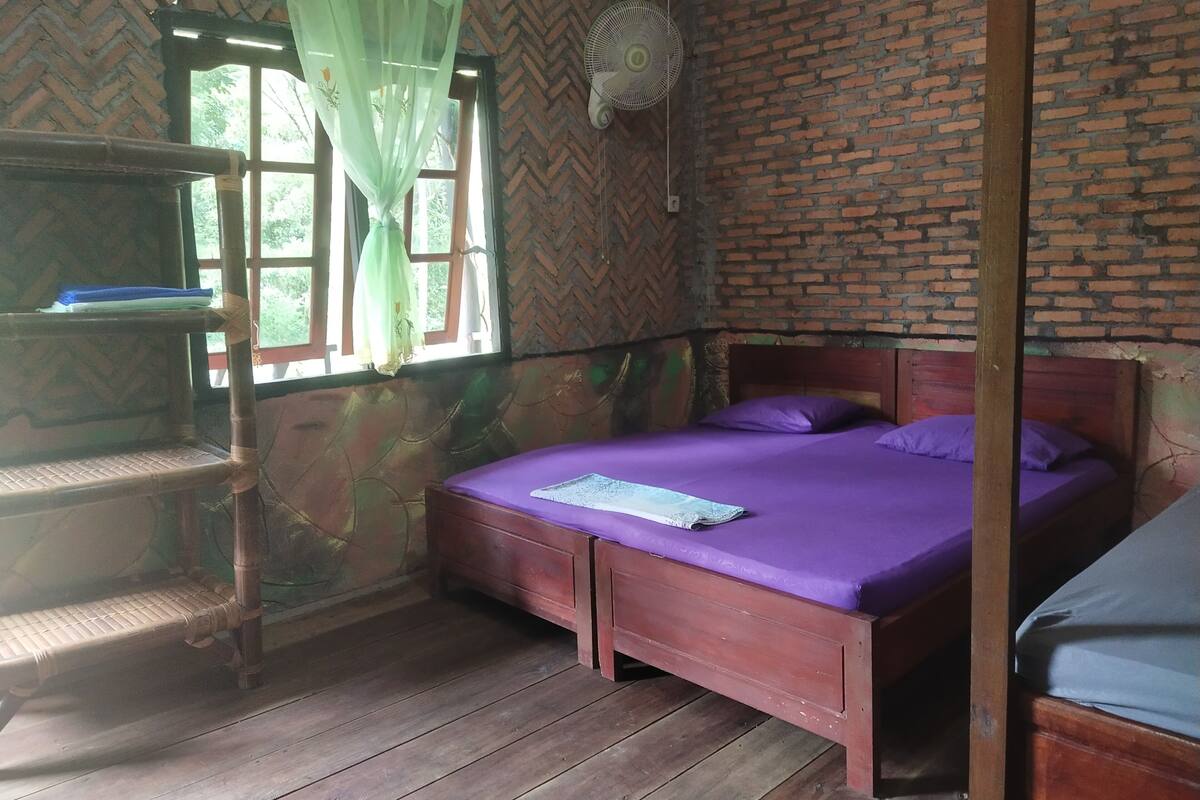 Een kamer met een bed, een plank, een raam met gordijn en een bakstenen muur.