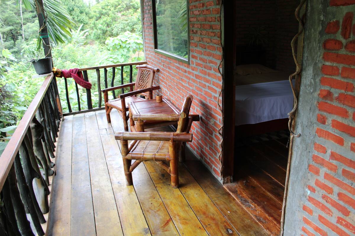 Een houten terras met een bed en een stoel, een bakstenen muur en uitzicht op groen.
