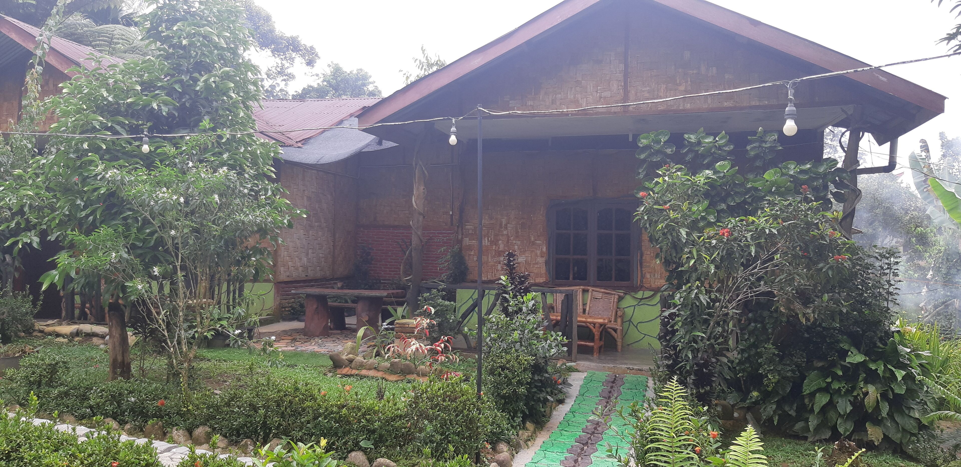 A rustic wooden house with a porch, surrounded by lush greenery and a garden.